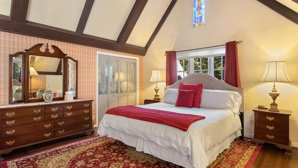 Cozy bedroom with a bed adorned in white and red linens, wooden furniture, and a stained glass window.